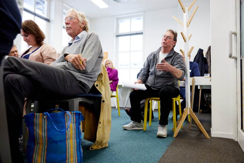 Twee deelnemers zitten op stoelen. Eén heeft een boodschappentas bij zich, de ander is aan het woord.